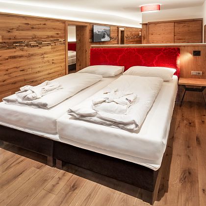 Gemütliches Doppelzimmer in einem modernen Hotel mit Holzelementen, roten Akzenten und Parkettboden. Zwei Bademäntel liegen auf dem Bett bereit.