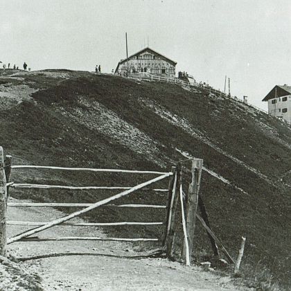 A black and white photo of a hillside with rustic buildings and a wooden gate in the foreground. The scene is set in a mountainous area.